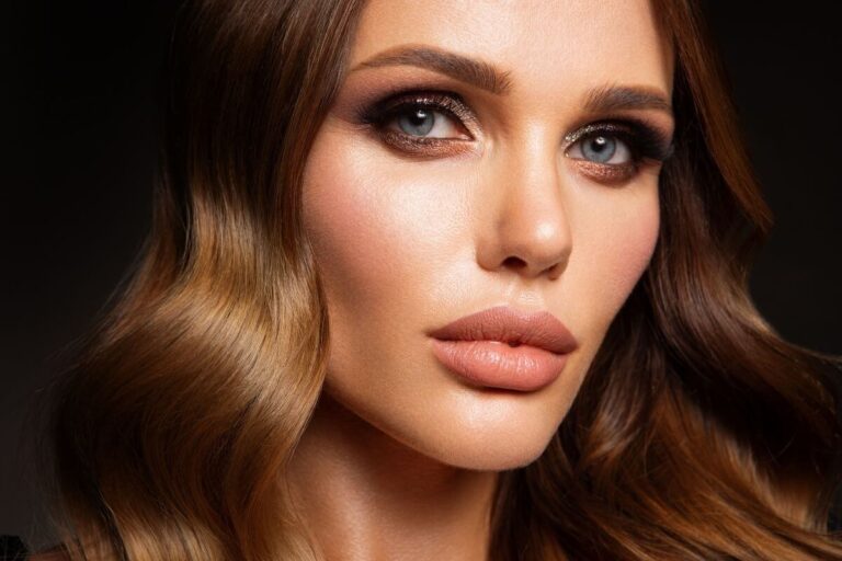 Woman with shiny, long, wavy brunette permanent hair colour and nude soft-glam makeup gazing into the camera.