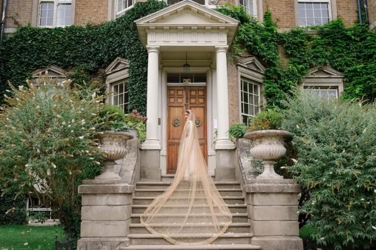 Bride in a wedding dress standing on stairs in front of Hampton Court House
